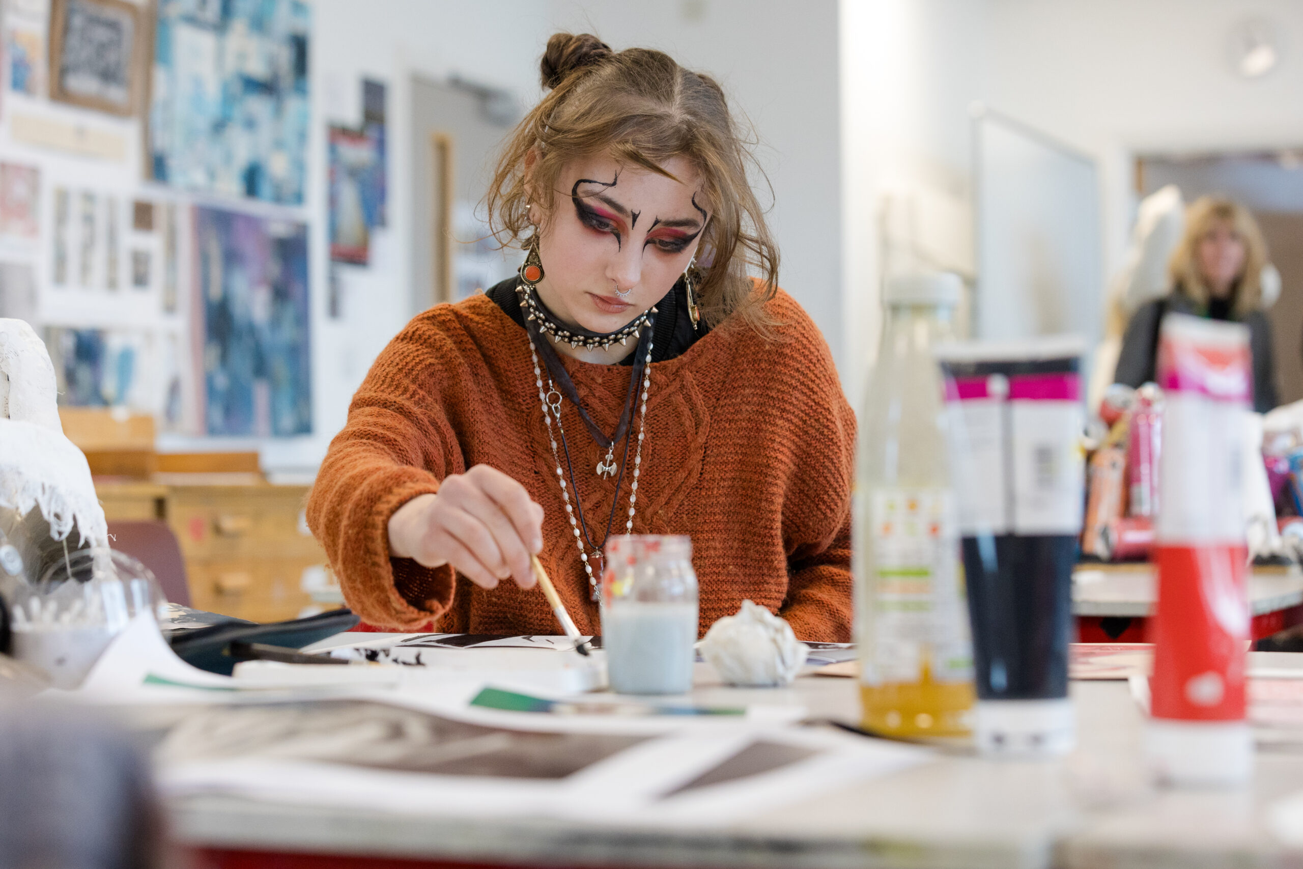 Person seated at a table engaged in painting, with art supplies including brushes, jars, and bottles spread across the workspace