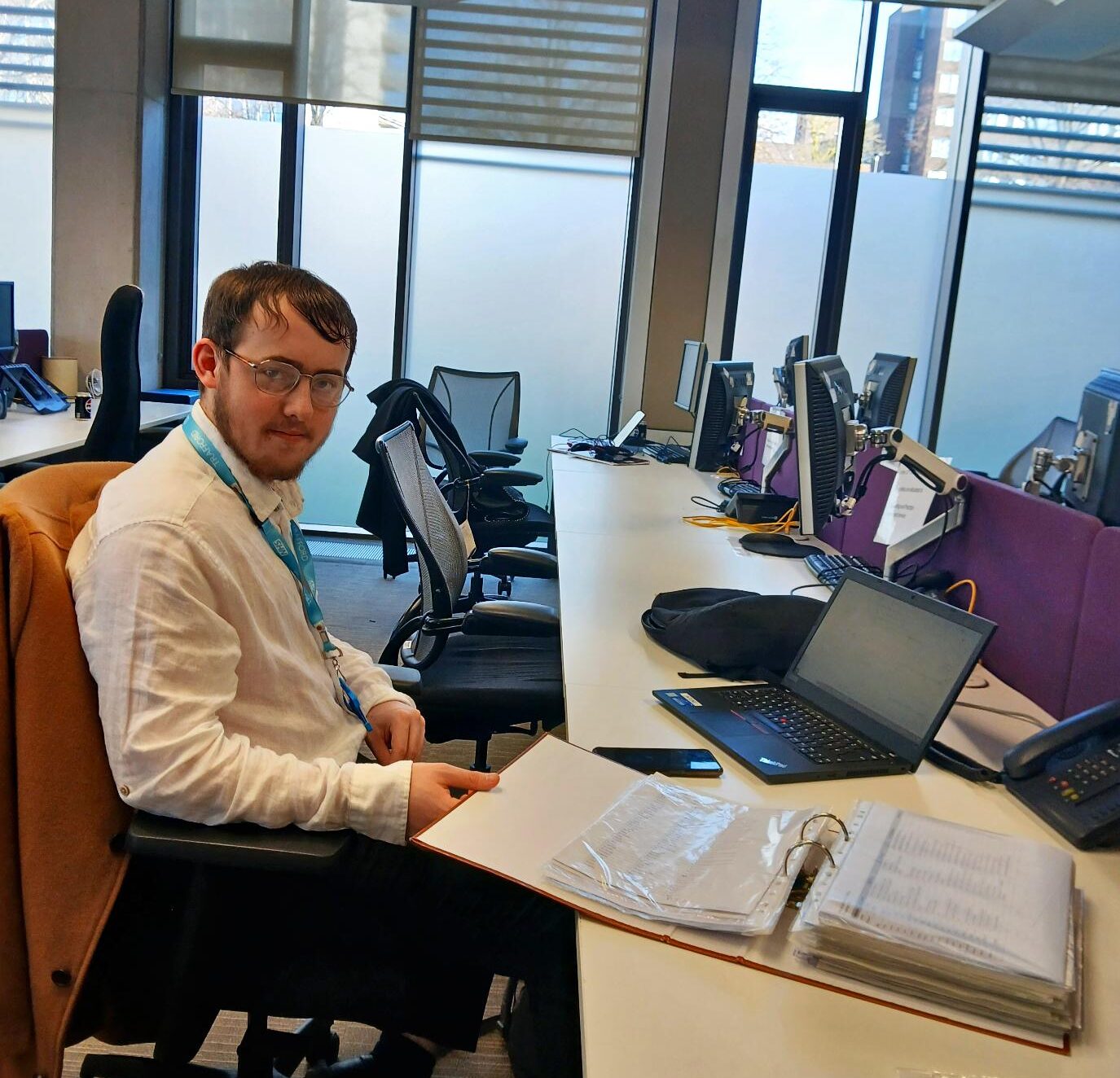 Boy seated at a desk in an office with multiple monitors, chairs, and documents.