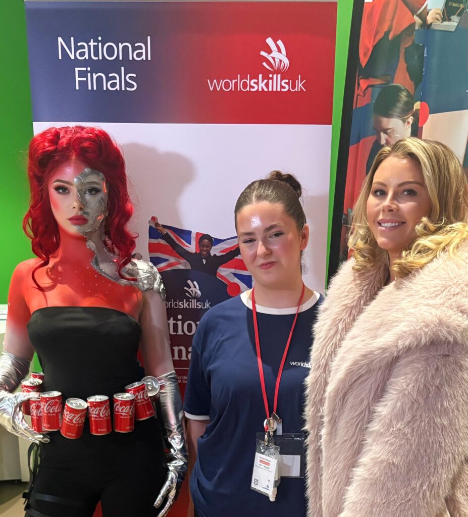 Three individuals standing in front of a WorldSkills UK banner that reads “National Finals.” One person is wearing an elaborate costume featuring metallic silver arm pieces and a belt made of Coca-Cola cans, with bright red hair. Another person is wearing a WorldSkills UK shirt with a lanyard and ID badge. The third person is dressed in a light-colored, fluffy coat. The backdrop includes the WorldSkills UK logo and an image of a person holding a Union Jack flag.