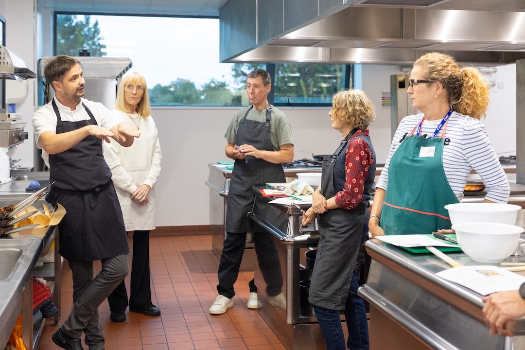 A tutor demonstrating a technique to a small group of adult learners in a professional training kitchen.